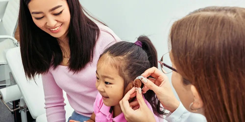 Child smiling as a clinician fits a hearing aid, with a parent observing, supporting the management of sensory hearing loss.