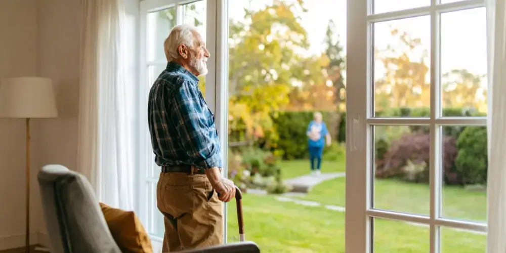 An elderly man with a walking cane stands comfortably in his home, looking out at his garden, thanks to a sustainable in-home care plan that supports his independence.